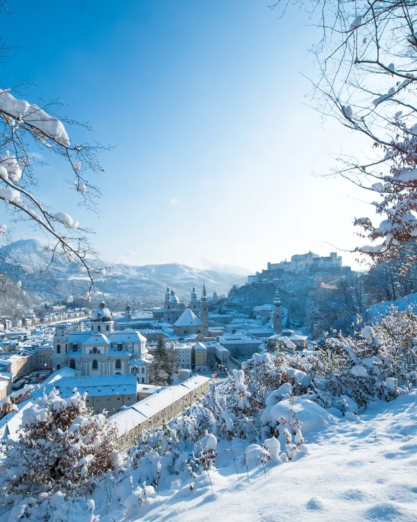 Winterliche Stadtansicht von Salzburg mit schneebedeckten Dächern und Festung Hohensalzburg im Sonnenlicht